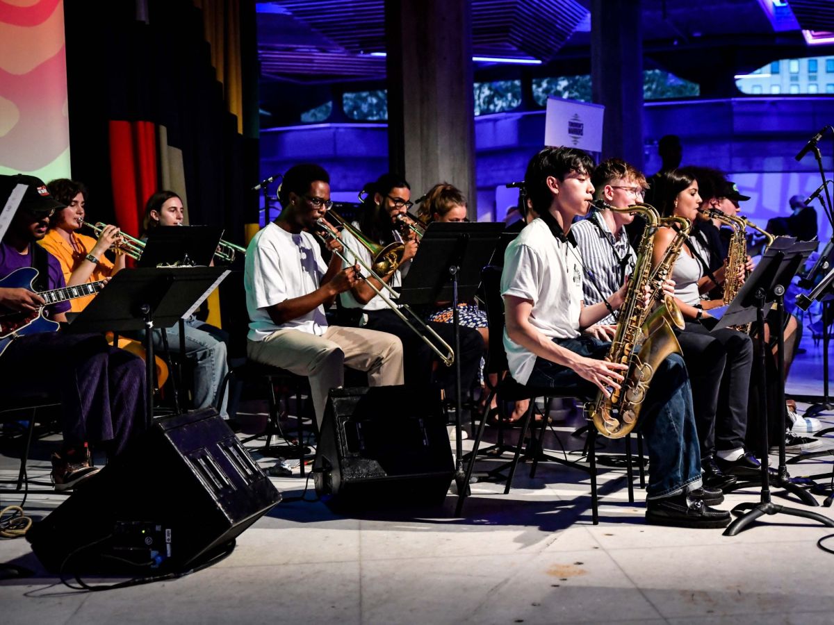 A photo showing more than 10 young people playing in an orchestra. They are all seated, and all but one are playing brass instruments, include saxophone and trombone.