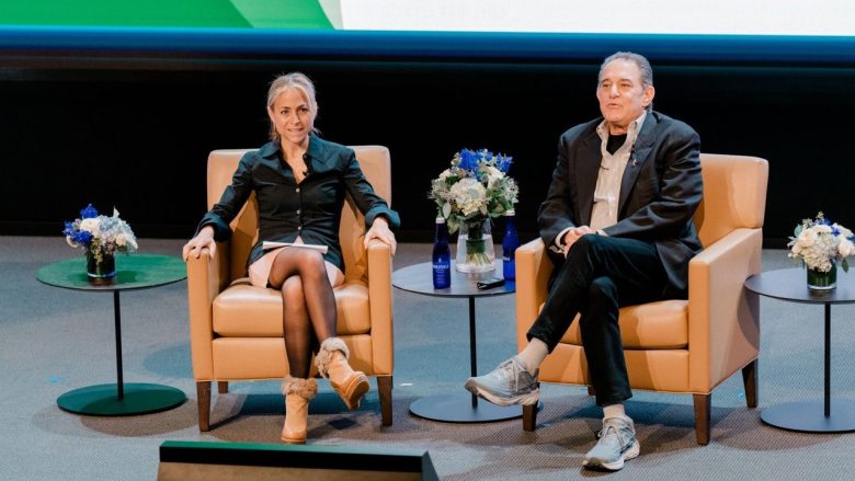 Cynthia Katz and an unidentified speaker seated on stage during a presentation, with floral arrangements and water bottles on the table.